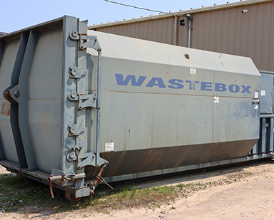 A large grey compactor outside of a commercial building.
