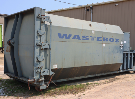 A large grey trash compactor that says "Waste Box" on the side located next to a brown commercial building.